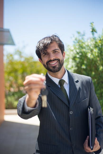 A smiling bearded real estate agent in a suit presenting a house key outdoors.