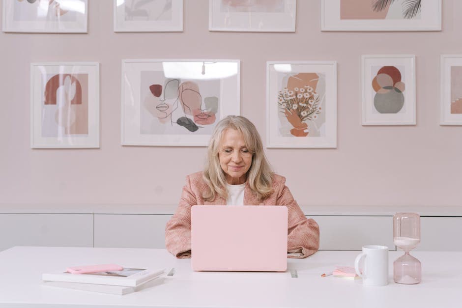 Elderly woman in pink coat working on a laptop at a white desk surrounded by artistic frames.