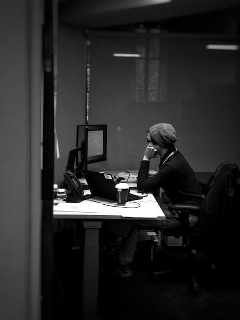 A man in a beanie works intently at a desk in an office. Black and white photography.