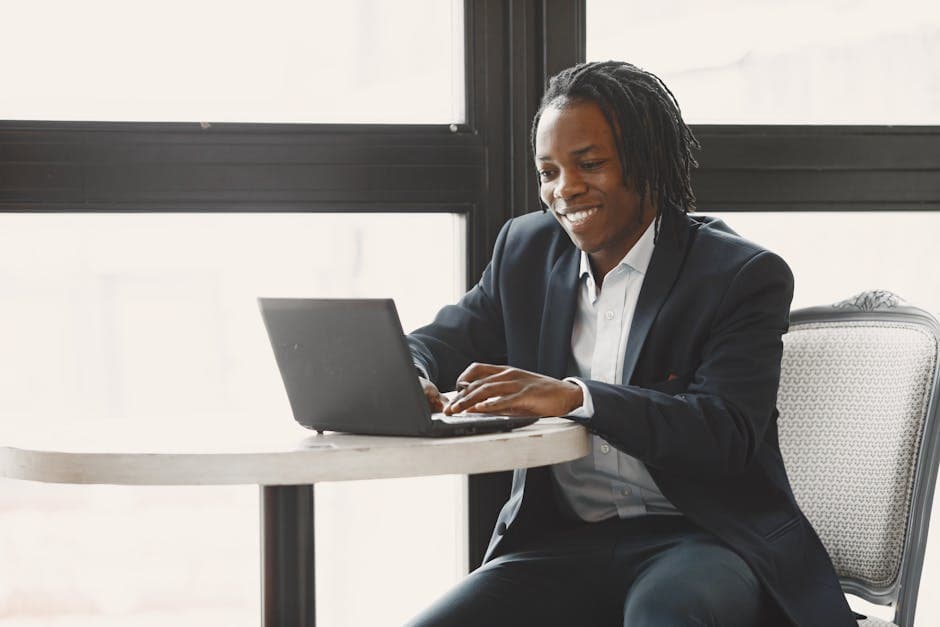 Smiling man in a suit working on a laptop at a table in a modern office setting.