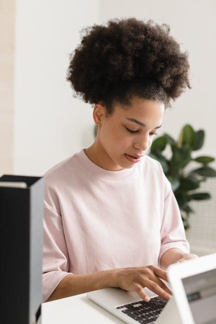 Young woman concentrating while typing on a laptop, seated at a desk indoors.