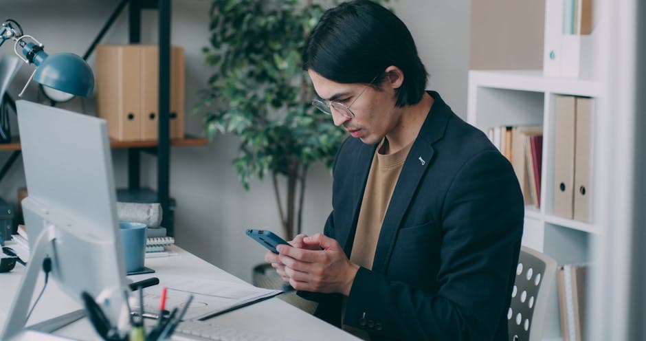 Young man in an office using smartphone while working at a computer desk.
