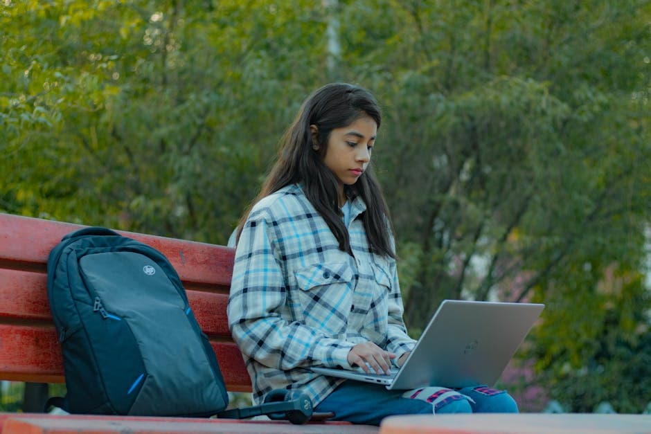 South Asian woman in plaid shirt using laptop on bench in park setting.