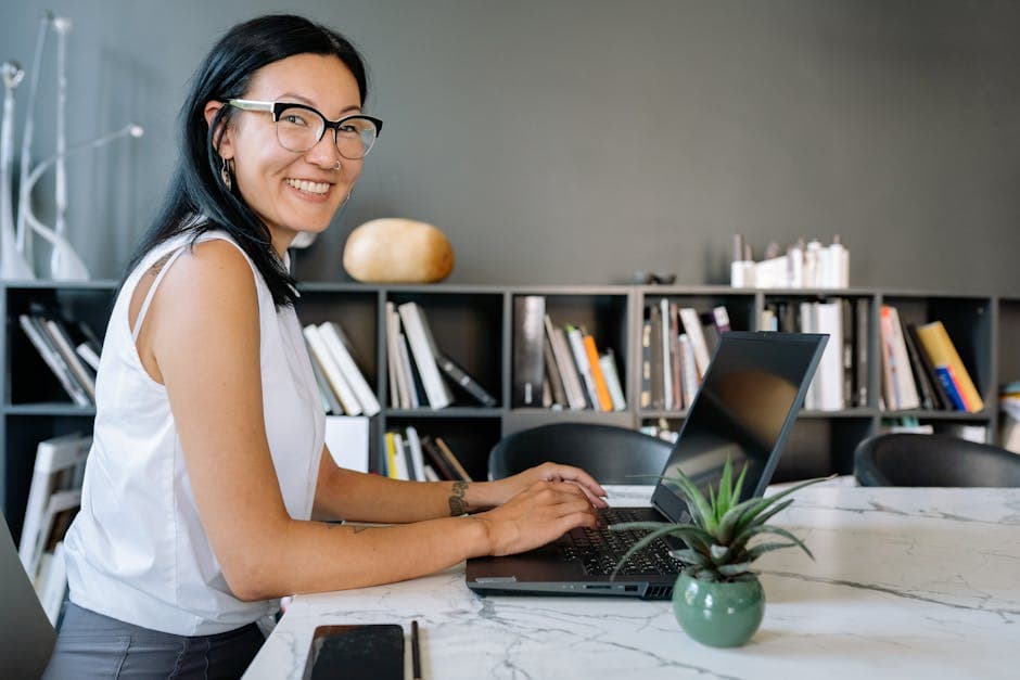 Cheerful businesswoman using laptop at a desk in a stylish office setup with bookshelves.