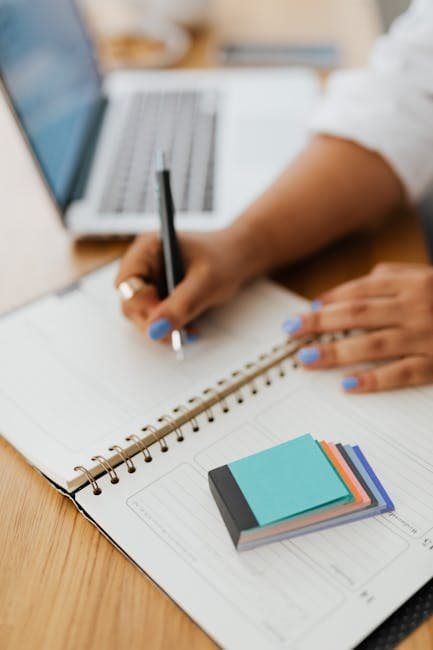Close-up of hands writing in notebook with laptop and sticky notes nearby.
