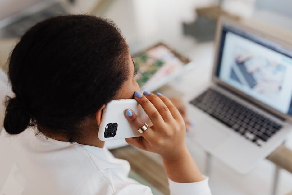 Woman holding smartphone to ear while working on a laptop. Modern communication and multitasking.