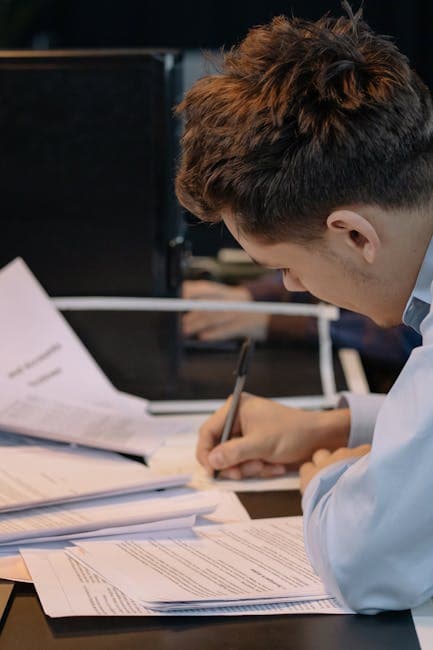 A young professional takes notes at a desk filled with documents in an office.