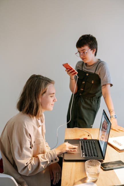 Two young professionals engaged in creative work using a laptop and smartphone in a bright, modern workspace.