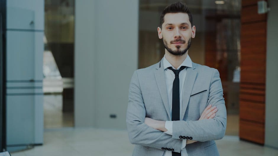 Businessman in a sleek gray suit with arms crossed, looking confident in a modern office.